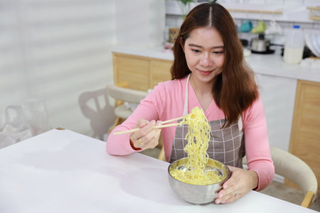 Portrait of young happy asian currly hair woman sitting on table and eating tasty and instant noodle for lunch in the kitchen with chopsticks and brick wall. Unhealthy and cheap food lifestyle conceptの写真素材