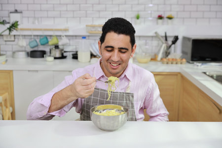 Closeup young happy hispanic currly hair man sitting on table and eating tasty and instant noodle for lunch in the kitchen with chopsticks and brick wall. Unhealthy and cheap food lifestyle conceptの写真素材