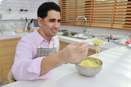 Closeup young happy hispanic currly hair man sitting on table and eating tasty and instant noodle for lunch in the kitchen with chopsticks and brick wall. Unhealthy and cheap food lifestyle conceptの写真素材