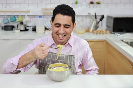 Closeup young happy hispanic currly hair man sitting on table and eating tasty and instant noodle for lunch in the kitchen with chopsticks and brick wall. Unhealthy and cheap food lifestyle conceptの写真素材