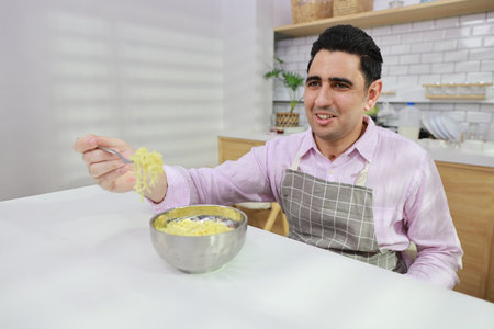 Closeup young happy hispanic currly hair man sitting on table and eating tasty and instant noodle for lunch in the kitchen with chopsticks and brick wall. Unhealthy and cheap food lifestyle conceptの写真素材