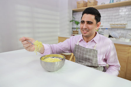 Closeup young happy hispanic currly hair man sitting on table and eating tasty and instant noodle for lunch in the kitchen with chopsticks and brick wall. Unhealthy and cheap food lifestyle conceptの写真素材