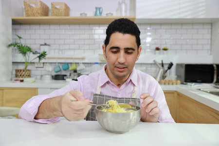 Closeup young happy hispanic currly hair man sitting on table and eating tasty and instant noodle for lunch in the kitchen with chopsticks and brick wall. Unhealthy and cheap food lifestyle conceptの写真素材