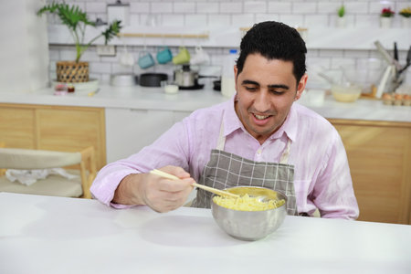 Closeup young happy hispanic currly hair man sitting on table and eating tasty and instant noodle for lunch in the kitchen with chopsticks and brick wall. Unhealthy and cheap food lifestyle conceptの写真素材