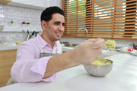 Closeup young happy hispanic currly hair man sitting on table and eating tasty and instant noodle for lunch in the kitchen with chopsticks and brick wall. Unhealthy and cheap food lifestyle conceptの写真素材