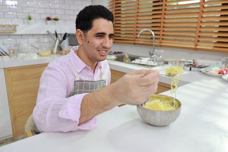 Closeup young happy hispanic currly hair man sitting on table and eating tasty and instant noodle for lunch in the kitchen with chopsticks and brick wall. Unhealthy and cheap food lifestyle conceptの写真素材