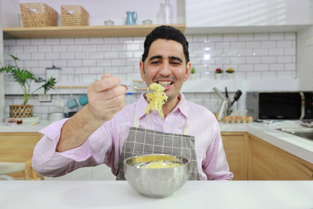 Closeup young happy hispanic currly hair man sitting on table and eating tasty and instant noodle for lunch in the kitchen with chopsticks and brick wall. Unhealthy and cheap food lifestyle conceptの写真素材