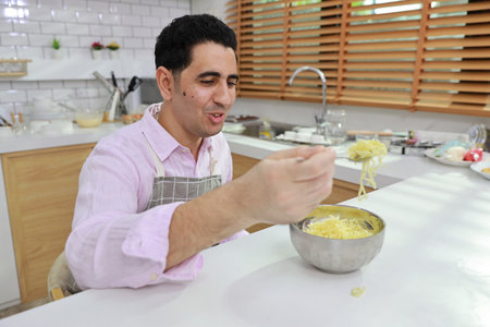 Closeup young happy hispanic currly hair man sitting on table and eating tasty and instant noodle for lunch in the kitchen with chopsticks and brick wall. Unhealthy and cheap food lifestyle conceptの写真素材
