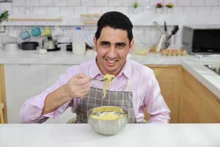 Closeup young happy hispanic currly hair man sitting on table and eating tasty and instant noodle for lunch in the kitchen with chopsticks and brick wall. Unhealthy and cheap food lifestyle conceptの写真素材