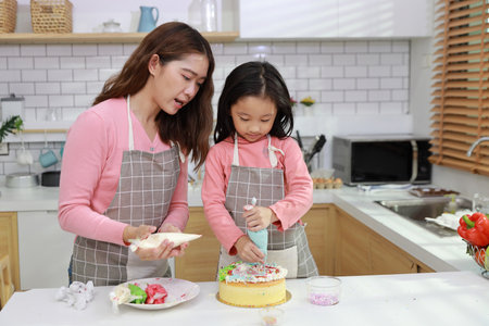 Happy lovely asian single mom and cute daughter in apron having fun with teaching and decorating homemade cake in the kitchen. Family lifestyle cooking with education concept.の写真素材