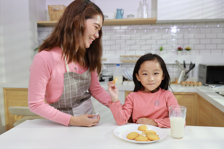 Kid asian black hair girl with mom sitting on table and drinking milk while eating vanilla cookies for breakfast with enjoying time. Tasty food and delicious food with happy meal lifestyle kid conceptの写真素材