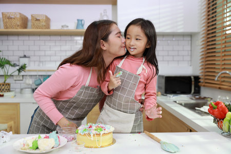 Happy lovely asian single mom and cute daughter in apron having fun with decorating homemade cake in the kitchen. Mother kiss daughter cheek. Family lifestyle cooking with education concept.の写真素材