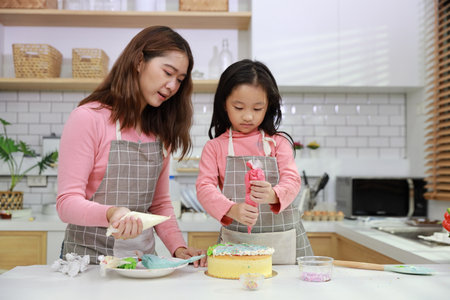 Happy lovely asian single mom and cute daughter in apron having fun with teaching and decorating homemade cake in the kitchen. Family lifestyle cooking with education concept.の写真素材