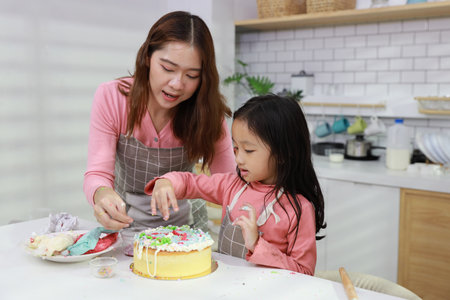 Happy lovely asian single mom and cute daughter in apron having fun with teaching and decorating homemade cake in the kitchen. Family lifestyle cooking with education concept.の写真素材
