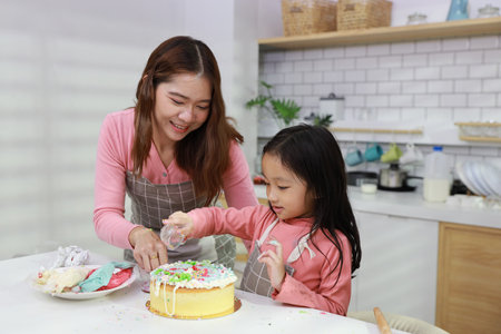 Happy lovely asian single mom and cute daughter in apron having fun with teaching and decorating homemade cake in the kitchen. Family lifestyle cooking with education concept.の写真素材