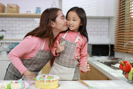 Happy lovely asian single mom and cute daughter in apron having fun with decorating homemade cake in the kitchen. Mother kiss daughter cheek. Family lifestyle cooking with education concept.の写真素材