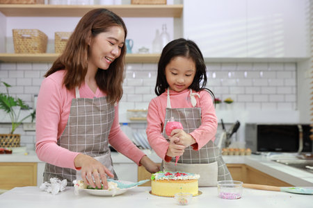Happy lovely asian single mom and cute daughter in apron having fun with teaching and decorating homemade cake in the kitchen. Family lifestyle cooking with education concept.の写真素材