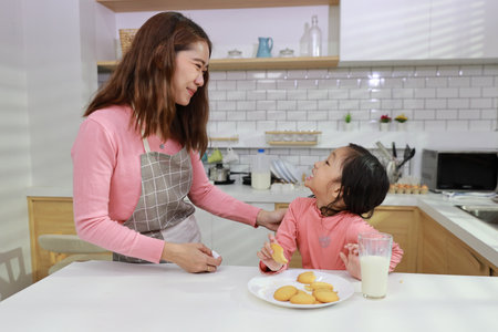Kid asian black hair girl with mom sitting on table and drinking milk while eating vanilla cookies for breakfast with enjoying time. Tasty food and delicious food with happy meal lifestyle kid conceptの写真素材