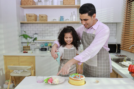 Happy lovely asian single dad and cute daughter in apron having fun with teaching and decorating homemade cake in the kitchen. Family lifestyle cooking with education concept.の写真素材