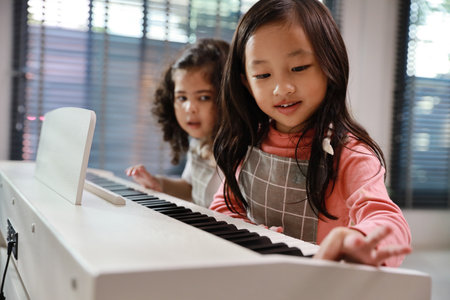 Kid asian black hair girl and hispanic currly hair girl sitting and playing piano in living room house indoor. Musical and relaxation makes them happiness. Health care lifestyle concept.の写真素材
