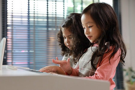 Kid asian black hair girl and hispanic currly hair girl sitting and playing piano in living room house indoor. Musical and relaxation makes them happiness. Health care lifestyle concept.の写真素材