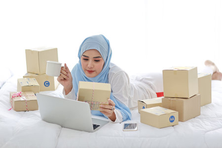Beautiful and young asian woman in muslim sleepwear with attractive look, sitting on bed with computer and online package box delivery. Startup small business SME freelance girl work with mobile phoneの写真素材