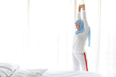 Beautiful asian woman wearing white muslim sleepwear, stretching her arms after getting up in the morning at sunrise. Cute young girl with blue hijab standing and relaxing with happy and smiling face.の写真素材