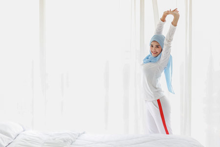 Beautiful asian woman wearing white muslim sleepwear, stretching her arms after getting up in the morning at sunrise. Cute young girl with blue hijab standing and relaxing with happy and smiling face.の写真素材