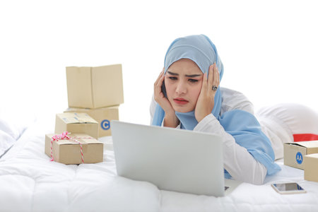 Beautiful and young asian woman in muslim sleepwear with headache emotion lies on bed with computer, mobile phone and online package box delivery. Smart girl with hijab receive bad news and tiredの写真素材