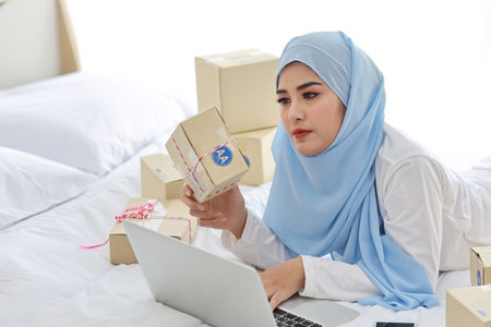 Beautiful and young asian woman in muslim sleepwear with attractive look, lies on bed with computer and online package box delivery. Startup small business SME freelance girl working with mobile phoneの写真素材