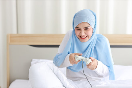 VDO game console station concept. active asian woman wearing white muslim sleepwear sitting on bed, holding joystick and playing exciting game. Cute girl looked excited with game controller consoleの写真素材