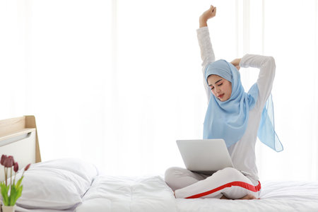 Young asian woman using computer sitting on bed in bedroom. Beautiful girl in muslim sleepwear with blue hijab stretching her arms after working before go to bed. Lifestyle and technology conceptの写真素材