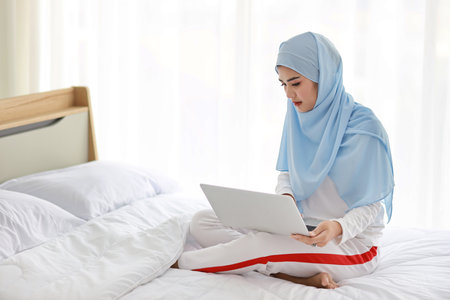 Young asian woman use laptop sitting on bed in bedroom. Beautiful girl in white muslim sleepwear with blue hijab check order or study from notebook before go to bed. Lifestyle and technology conceptの写真素材
