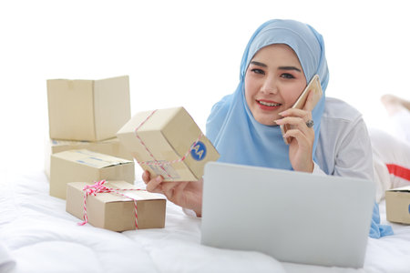 Beautiful and young asian woman in muslim sleepwear with attractive look, lies on bed with computer and online package box delivery. Startup small business SME freelance girl working with mobile phoneの写真素材
