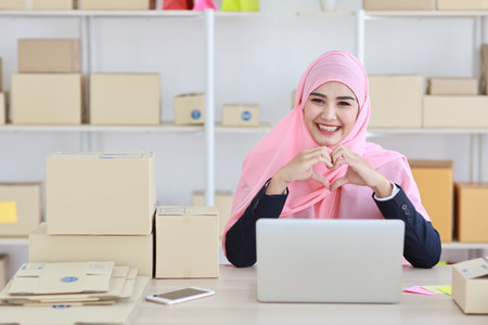 Active asian woman in blue muslim suit sitting and showing love symbole while working with computer and online package box delivery. Startup small business freelance working girl conceptの写真素材