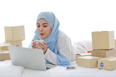 Young attractive asian woman in sleepwear muslim dress, holding coffee cup on bed with computer and online package box delivery. Startup small business SME freelance girl working with mobile phoneの写真素材
