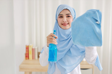 Indoor shot active young beautiful asian muslim housewife woman holding cleaning spray, washing window glass. Portrait cute girl with happy emotional cleaning bedroom in new apartment after moved in.の写真素材