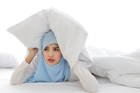 Oversleeping asian woman in muslim sleepwear lying on bed and missing ring of alarm clock waking up. Young girl trying to sleep by putting pillow over her head in bedroom with unhappy emotionの写真素材