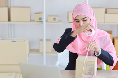 Active smiling asian woman in blue muslim suit sitting and working with online package box delivery. Startup small business SME freelance girl working on computer and mobile phone with happy face.の写真素材