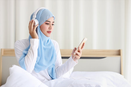 Portrait of beautiful asian female in muslim sleepwear watching online story on mobile phone, lites on bed and connected with wireless internet. Young cute girl with hijab listen music from smartphoneの写真素材