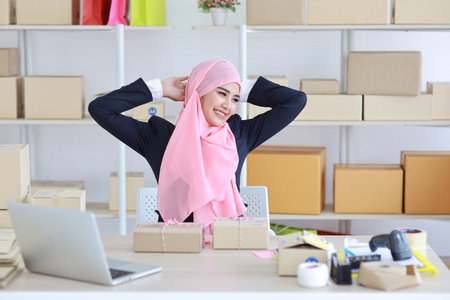 Active asian woman in blue muslim suit sitting and stretching after working with computer and online package box delivery. Startup small business freelance working girl conceptの写真素材