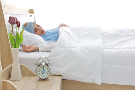Oversleeping asian woman wearing white muslim sleepwear lying on bed, missing ring of alarm clock waking up. Side view portrait young girl hand trying to stop ringing while her hand on her faceの写真素材