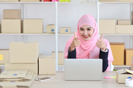 Active asian woman in blue muslim suit sitting and working with computer and online package box delivery. Startup small business freelance working girl showing thumb up with exciting emotion.の写真素材