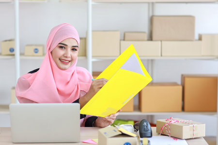 Active asian woman wearing muslim suit sitting and holding folder with computer and online package box delivery. Startup small business freelance concept.の写真素材