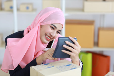 Active smiling asian woman in blue muslim suit sitting and working with online package box delivery. Startup small business SME freelance girl working on computer and mobile phone with happy face.の写真素材