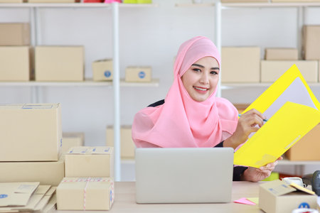 Active asian woman wearing muslim suit sitting and holding folder with computer and online package box delivery. Startup small business freelance concept.の写真素材