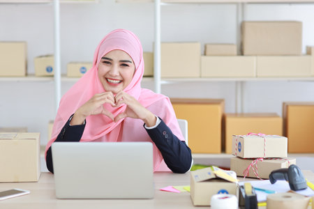 Active asian woman in blue muslim suit sitting and working with computer and online package box delivery. Startup small business freelance working girl showing love symbol with exciting emotion.の写真素材