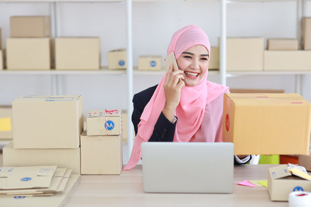 Religious asian woman in muslim dress sitting and using smart mobile phone and talking to customer with happy face and business package SME box delivery background.の写真素材