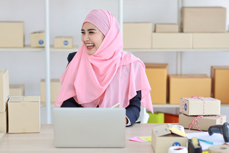 Religious asian woman in muslim dress sitting and using smart mobile phone and talking to customer with happy face and business package SME box delivery background.の写真素材