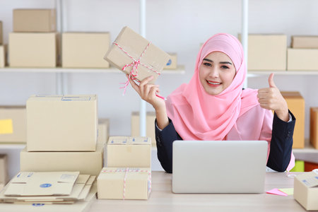 Active asian woman in blue muslim suit working with computer and showing thumb up and online package box delivery with happy smiling face. Startup small business freelance working girl look at productの写真素材